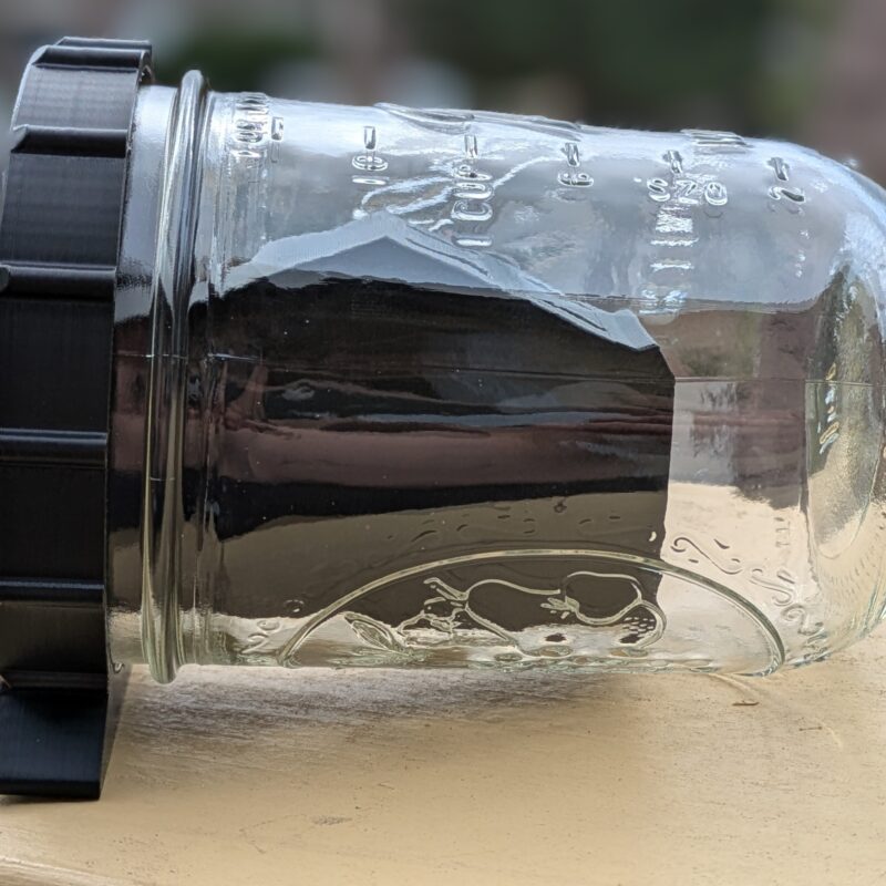 Mason jar mousetrap with a black CatchCap lid resting horizontally on a wooden railing outdoors.